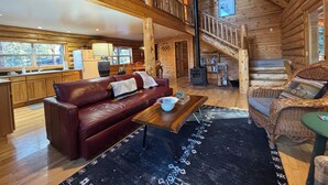 Een ruim houten blokhut interieur met een hoge plafond, een houten trap, een leren bank, een eethoek en een open haard.