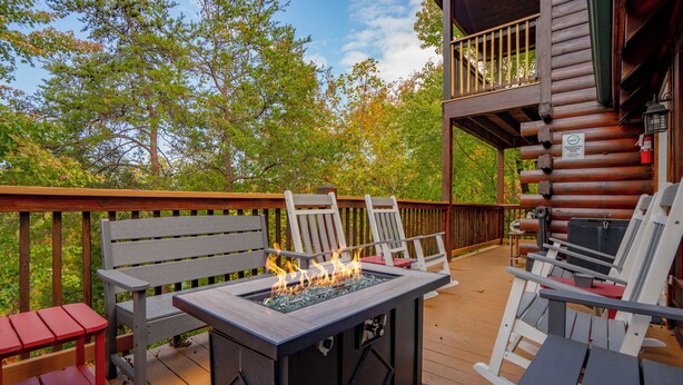A wooden deck with outdoor seating, a fire feature, and a view of trees. - SmokyMountainHigh: Pets, Hot Tub, Wifi, Resort Pool, Theater, Games, Fire Pit (Pigeon Forge)