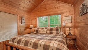 A wooden cabin bedroom with a plaid bedspread, two bedside lamps, and a window with blinds. - SmokyMountainHigh: Pets, Hot Tub, Wifi, Resort Pool, Theater, Games, Fire Pit (Pigeon Forge)