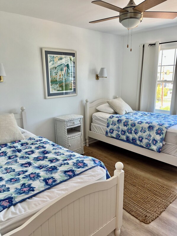 A bedroom with two beds, a ceiling fan, a framed picture, a bedside lamp, and a window with curtains. - Cape May Vacation Rentals-1 block to beach & 2 to town! Walk everywhere! (Cape May)