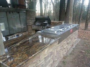 An outdoor kitchen with a stone wall, a gas stove, and a grill. - Large Modern Cabin with Hot Tub Tucked in Woods (Swan Lake)