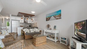Ein modernes Wohnzimmer mit einer Couch, Korbstühlen, einem Glastisch, einer weißen TV-Konsole und einer Küche im Hintergrund.