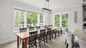 A spacious dining area with a long wooden table and chairs, large windows with a view of greenery, and a kitchen area visible to the right. - Luxury Lakefront Estate on Lake Anna 8,000 SqFt, Private Beach & Dock, Sleeps 20 (Bumpass)