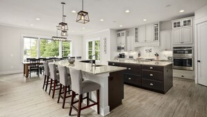A modern kitchen with white cabinets, a large island with bar stools, and a view of the outdoors through large windows. - Luxury Lakefront Estate on Lake Anna 8,000 SqFt, Private Beach & Dock, Sleeps 20 (Bumpass)