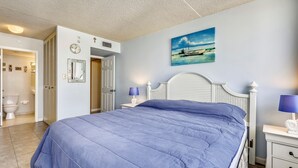 A bedroom with a bed, a nightstand, a lamp, a painting, and a bathroom visible through an open door.