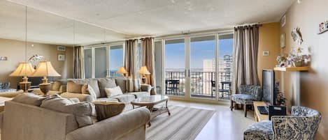 Amplia sala de estar con un sofá grande, una mesa de centro y un televisor. Hay cortinas en las ventanas, y se ve un balcón a través de las puertas de vidrio.