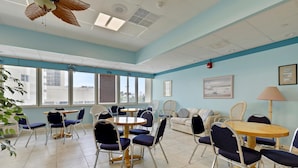 A room with multiple seating arrangements including tables and chairs, large windows, and a ceiling fan.