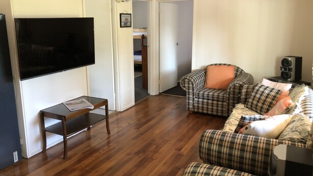 A living room with a flat-screen TV, plaid furniture, a small table, and a door leading to a bedroom. - Plough Cottage is a charming and cozy getaway for the family at a cheap price (Howqua)