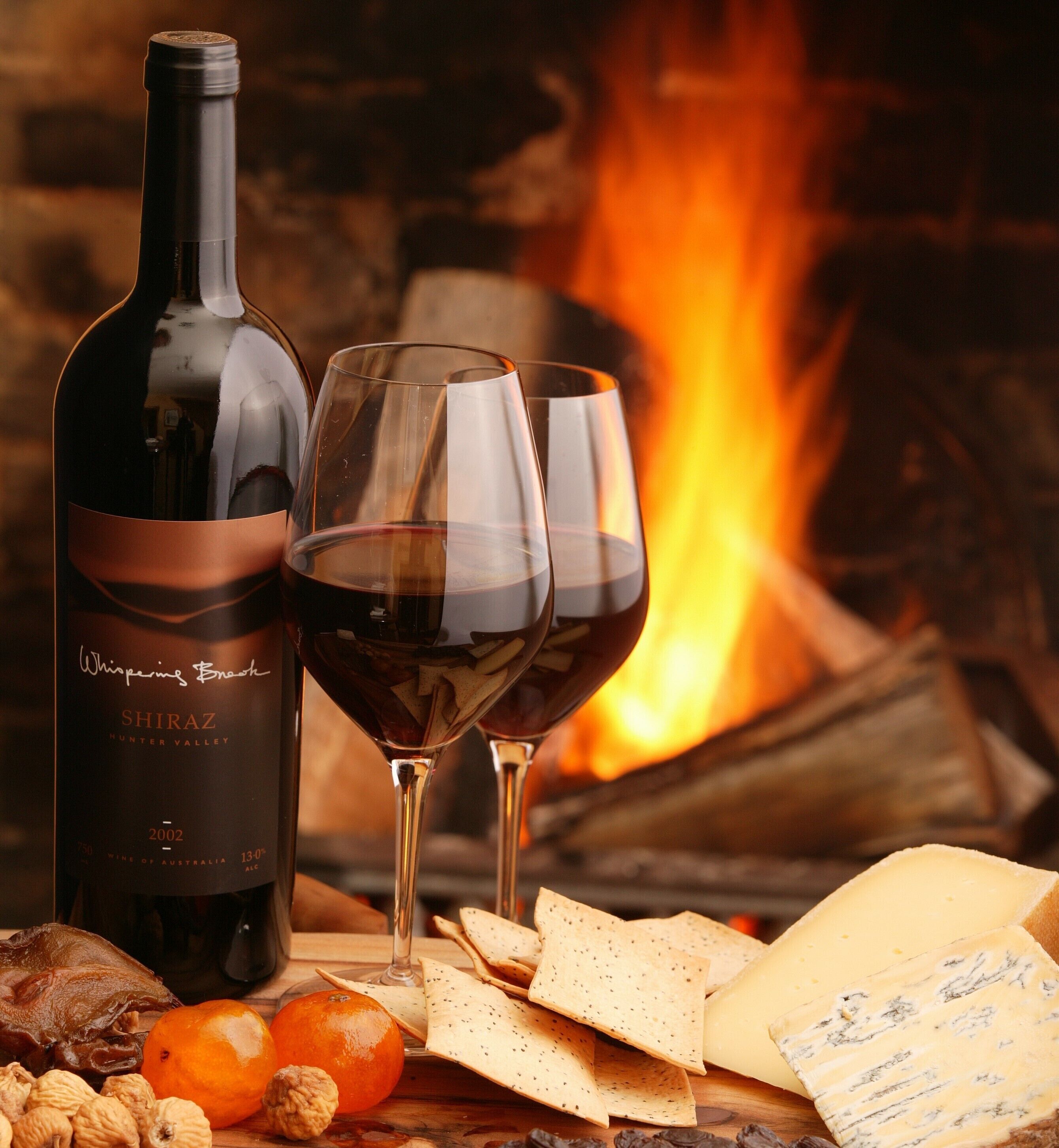 Une bouteille de vin Shiraz, deux verres à vin, des crackers, du fromage, des noix et des oranges sur une table en bois, avec une cheminée en arrière-plan.