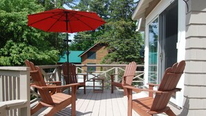 Une terrasse en bois, avec des fauteuils Adirondack et un parasol rouge, donnant sur une maison dont le toit est vert.