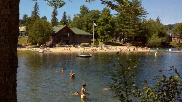 Une scène au bord d’un lac, avec un chalet en bois, une jetée, et des personnes qui nagent et se détendent sur la rive.