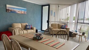 Een moderne woonkamer met een eethoek, voorzien van een groot raam met zonwering, een schilderij aan de muur en uitzicht op de omgeving.