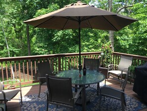 Une table ronde en verre, quatre chaises et un grand parasol sur une terrasse en bois, le tout entouré de verdure.