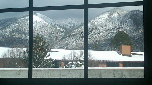 Une vue à travers une fenêtre sur un paysage montagneux enneigé, avec un arbre et un bâtiment doté d’un toit plat.