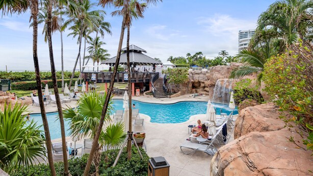A swimming pool surrounded by palm trees and lounge chairs, with a waterfall feature and a covered area with stairs. - Direct Ocean Front Property (Miami Beach)