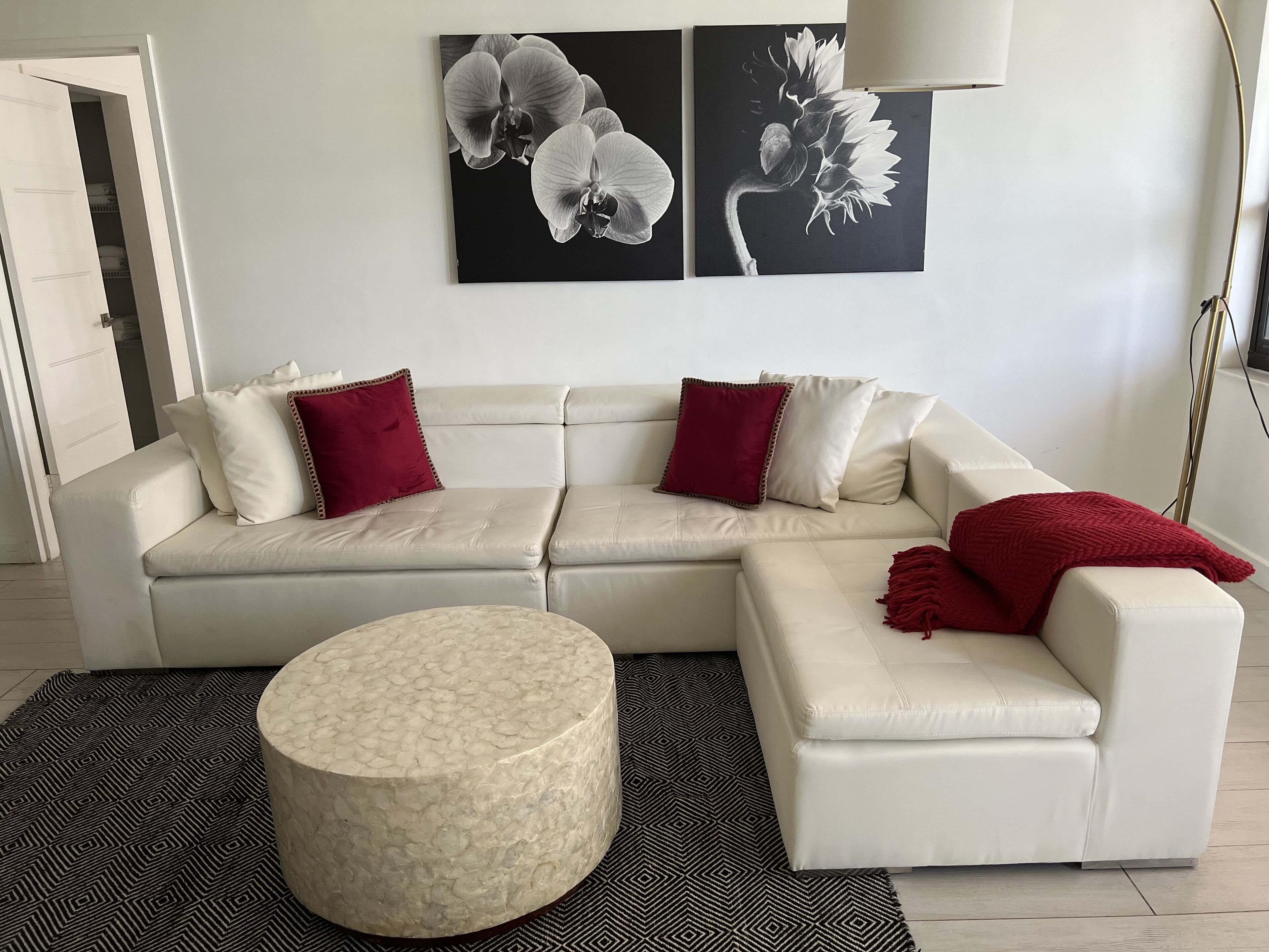En moderne stue med en beige hjørnesofa, to røde pynsepuder, en rund fodsæde og et sort-hvidt gulvtæppe med et mønster.