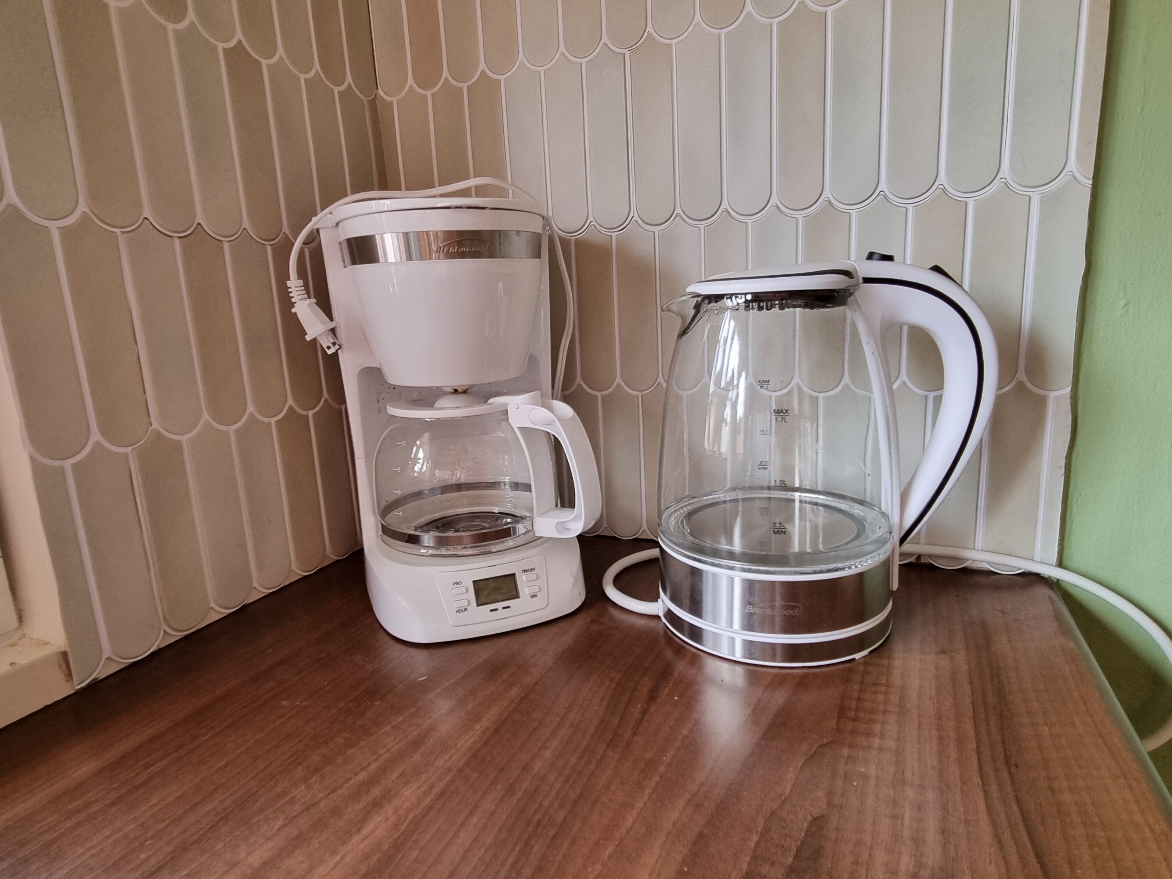 A coffee maker and a glass carafe on a wooden table against a tiled wall.