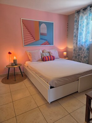 Un dormitorio con cama, mesita de noche, lámpara, un cuadro en la pared y ventana con cortinas.