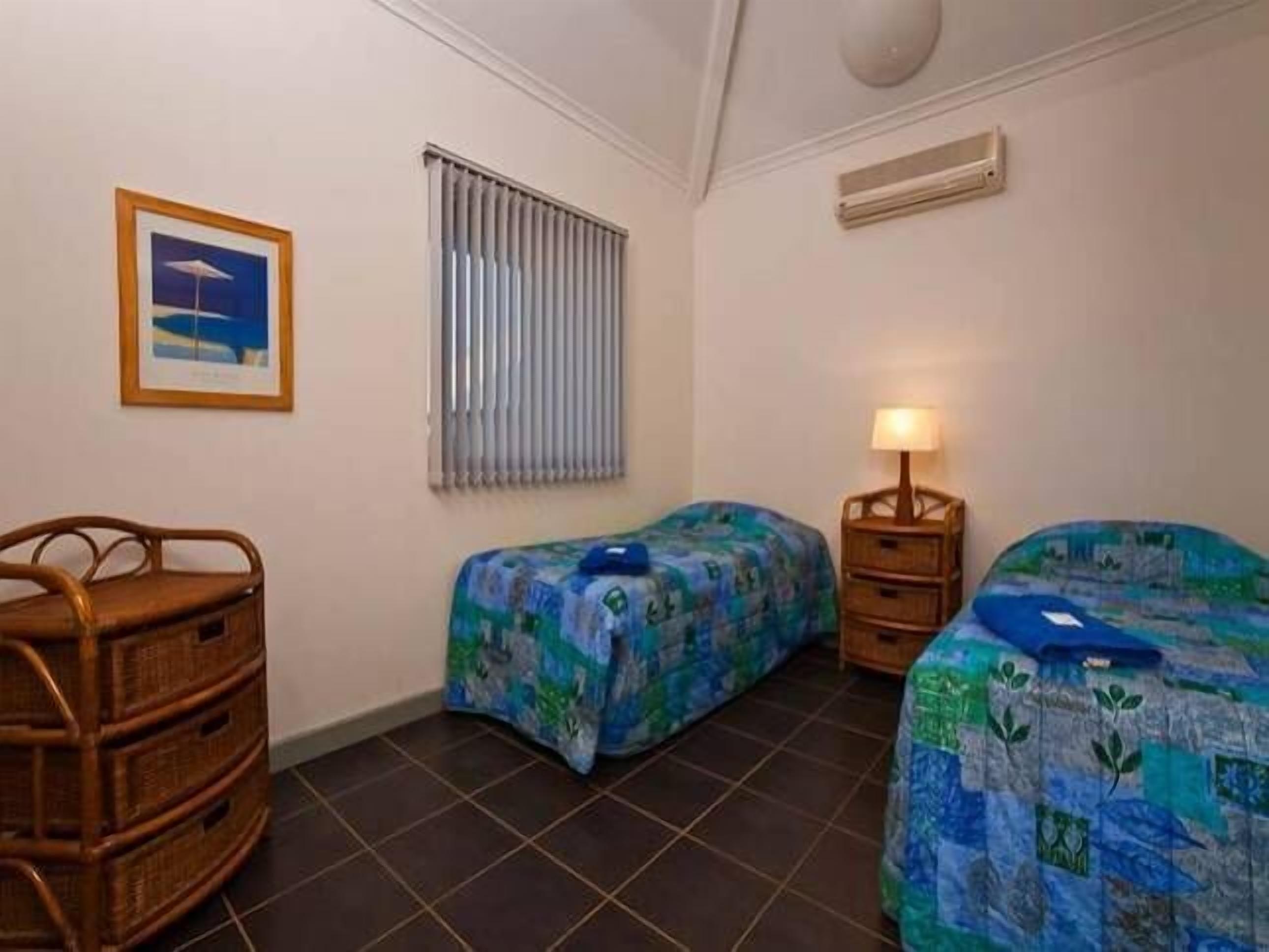 A room with two beds, a wicker chair, a nightstand with a lamp, a framed picture on the wall, and a window with blinds.