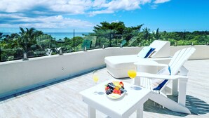 Una terraza en la azotea con sillones blancos, una mesa con una bandeja de frutas y bebidas, y vistas al océano y al verdor.