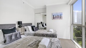 Ein Hotelzimmer mit zwei Betten, einem großen Fenster mit Blick auf Gebäude und Grünflächen, einem Gemälde an der Wand und einem Spiegel, der den Raum reflektiert.