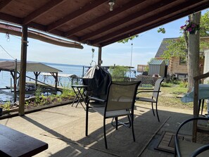 An outdoor patio with a covered seating area overlooking a body of water, featuring a grill and a view of a wooden cabin.