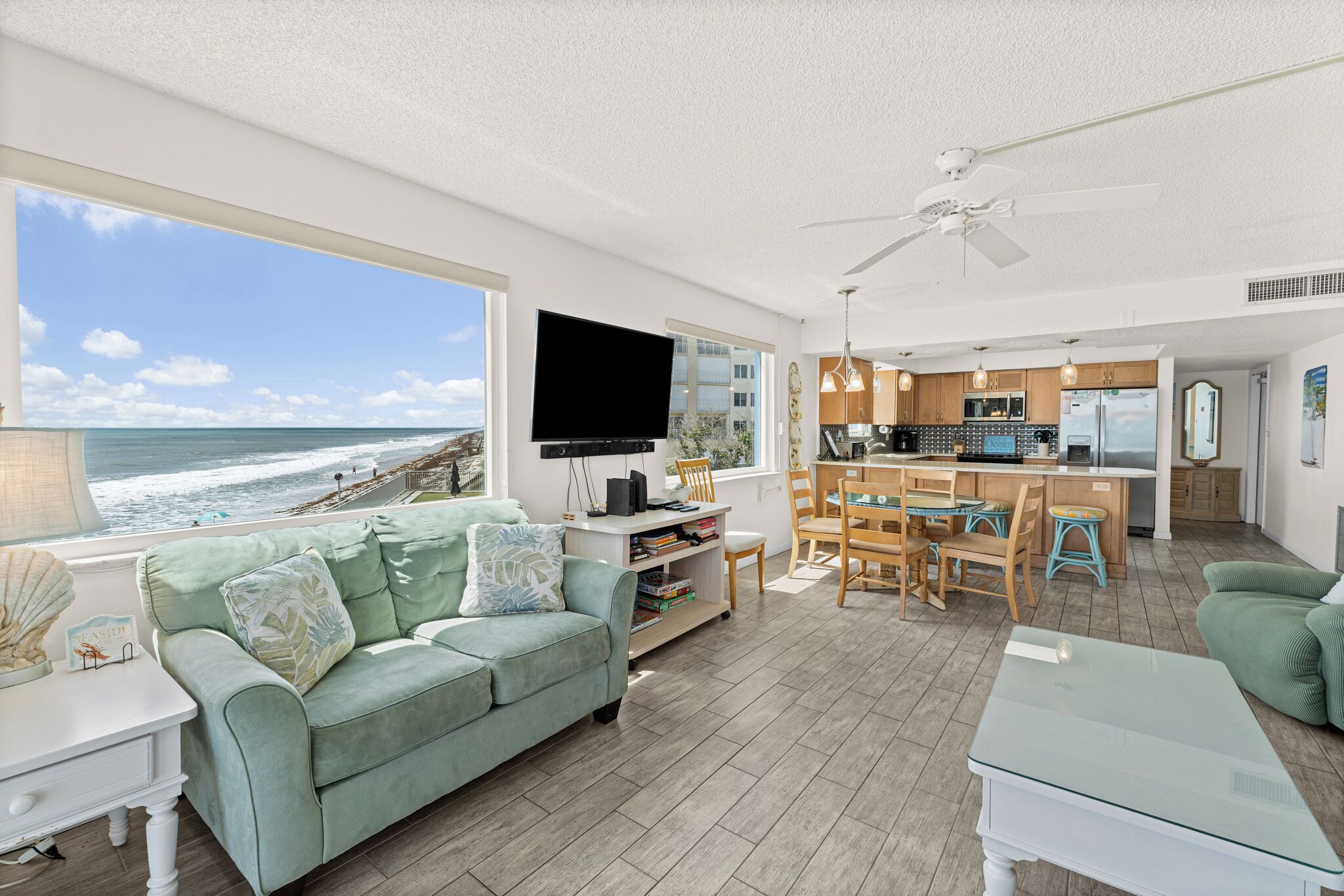 A living room with a large window overlooking the ocean, a flat-screen TV, and a kitchen area with wooden cabinets and a dining set.