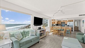 A living room with a large window overlooking the ocean, a flat-screen TV, and a kitchen area with wooden cabinets and a dining set. - Oceanfront 2BR Condo w/ Balcony & Panoramic Atlantic Views, Unit 226 (Satellite Beach)