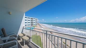 A balcony with a view of the beach and ocean, a pool, and a building with multiple floors.
