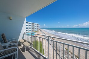 A balcony with a view of the beach and ocean, a pool, and a building with multiple floors.