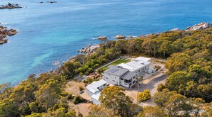 A modern two-story house with a flat roof, surrounded by dense green trees and a clear blue sea.