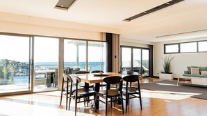 Un comedor moderno con una mesa grande y sillas, ventanas amplias con vista al mar, y una zona de estar acogedora con un sofá y una planta.
