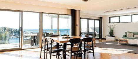 A modern dining area with a large table and chairs, large windows with a view of the sea, and a cozy seating area with a sofa and a plant.