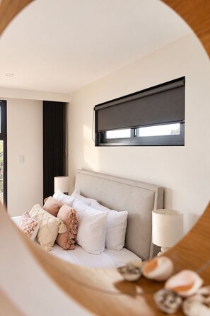 Un dormitorio con cama, almohadas, lámpara de noche, ventana con persiana y vistas al exterior.
