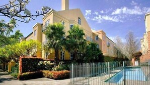 Un edificio de varios pisos con piscina, rodeado por una cerca metálica y varios árboles.