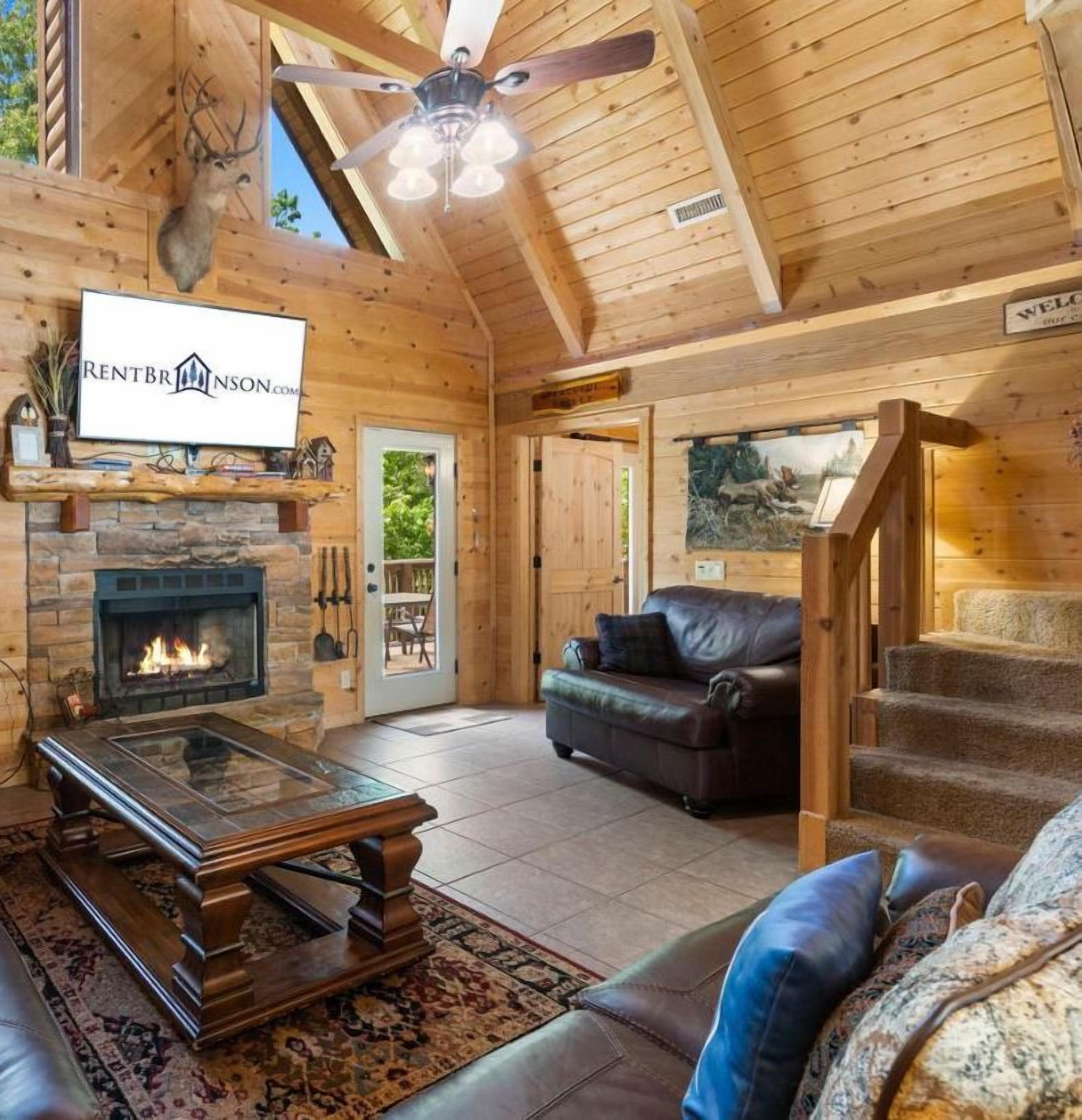A log cabin interior with a fireplace, leather furniture, and a television displaying 'RENTBRINSON.COM'.