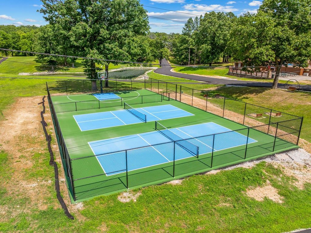 Un court de tennis extérieur doté d’une surface verte et de lignes bleues, entouré d’une clôture et d’arbres.