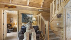 L’intérieur d’un chalet chaleureux, avec un grand ours en peluche sur un canapé en cuir, des murs en bois, un escalier et une pancarte de bienvenue.