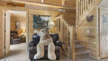 A cozy cabin interior with a large teddy bear on a leather couch, wooden walls, a staircase, and a welcome sign.