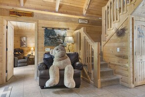 L’intérieur d’un chalet chaleureux, avec un grand ours en peluche sur un canapé en cuir, des murs en bois, un escalier et une pancarte de bienvenue.