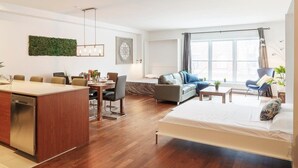 Una habitación de hotel moderna con suelo de madera, una cama, un sofá, zona de comedor y un ventanal grande.