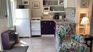 A compact kitchen with a refrigerator, stove, microwave, and cabinets. There is a colorful chair and a lamp on a side table. - Come visit in 2027! (Mt. Desert)