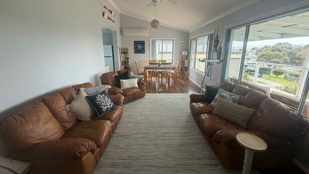 A spacious living room with leather sofas, a dining table, and large windows offering a view of the outdoors. - 63 Vista Drive, Mallacoota (Mallacoota)