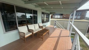 A covered outdoor area with a wooden bench and a sofa, large windows, and a view of a residential area. - 63 Vista Drive, Mallacoota (Mallacoota)