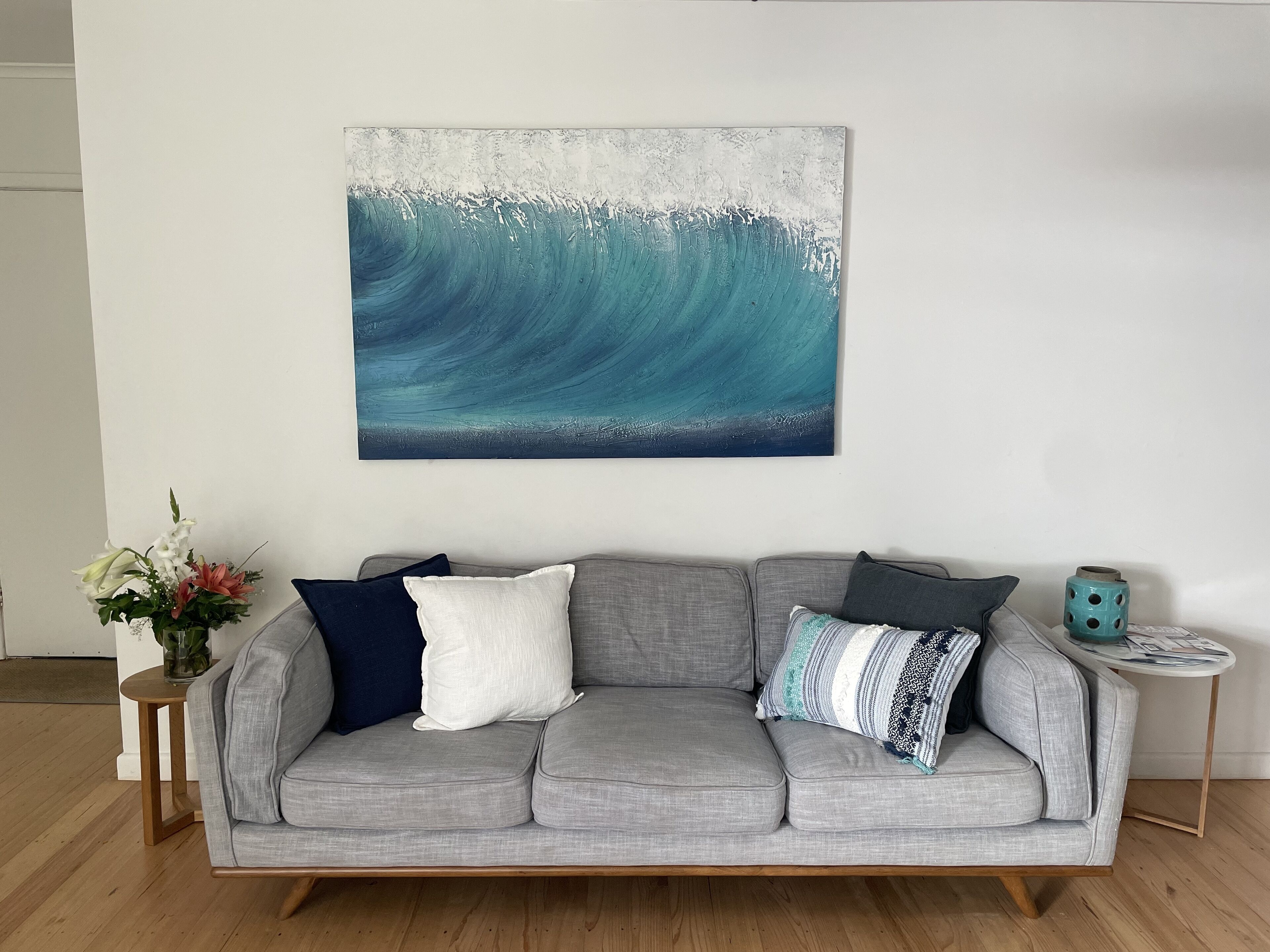 Ein graues Sofa mit mehreren Kissen, ein kleiner runder Tisch mit einer türkisfarbenen Vase und eine Blumenkomposition auf einem Beistelltisch.