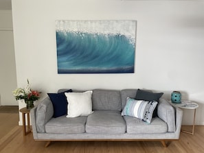 A grey sofa with multiple cushions, a small round table with a teal vase, and a floral arrangement on a side table.