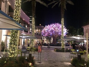 Un espacio al aire libre con mesas y sillas, un árbol grande iluminado y un edificio con balcones al fondo.