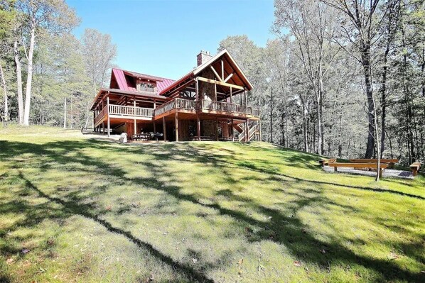 A large wooden cabin with a balcony and a covered porch, surrounded by a forested area.