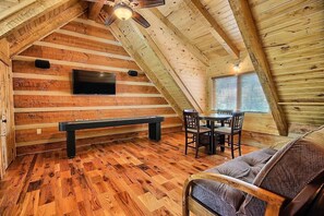 A cozy attic room with wooden walls, a bench, a table with chairs, and a TV mounted on the wall.