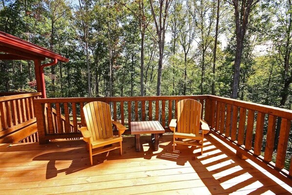 Eine Holzterrasse mit zwei Adirondack-Stühlen und einem kleinen Tisch, umgeben von einem Holgitter, mit Blick auf eine bewaldete Landschaft.